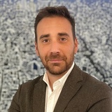 A professional man in a suit jacket smiling in front of an abstract cityscape painting.