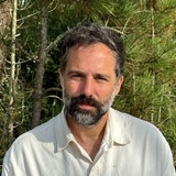 A man with a beard stands outdoors with trees in the background.