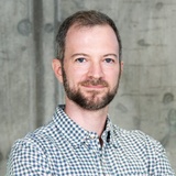 A man with a beard, wearing a checkered shirt, smiles gently in a professional setting.