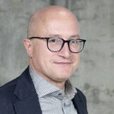 A professional portrait of a bald man wearing glasses, dressed in a formal suit and a checkered shirt.