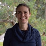 A smiling woman with brown hair wearing a blue scarf outdoors with trees in the background.