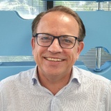 A smiling man wearing glasses and a light blue checkered shirt in a office environment.