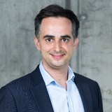 A professional portrait of a smiling man wearing a suit jacket and a blue shirt against a textured gray background.
