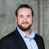 A professional headshot of a smiling bearded man wearing a suit jacket and a blue shirt.