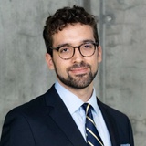 A professional portrait of a man in a suit and glasses, smiling at the camera.