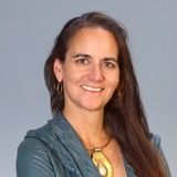 Portrait of a smiling woman with dark hair, wearing a teal blouse and a large golden necklace.