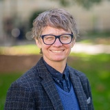 A portrait of a smiling woman with short hair wearing glasses, a blazer, and a collared shirt, standing outdoors.