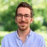 A portrait of a smiling man with glasses wearing a blue shirt in a park setting.