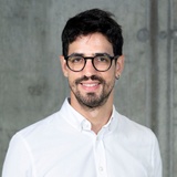 A portrait of a smiling man wearing glasses and a white shirt against a blurred background.