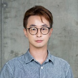 Portrait of a man with glasses wearing a striped shirt, standing in front of a concrete wall.
