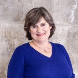 A cheerful mature woman in a blue top poses against a concrete background.