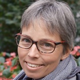 A portrait of a smiling woman with short grey hair wearing red and black glasses, standing outdoors.