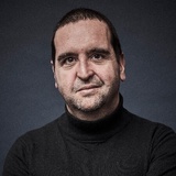A portrait of a middle-aged man with a stubble wearing a black turtleneck, looking directly at the camera.