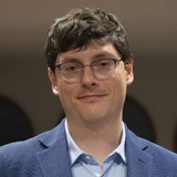 A young man with dark hair wearing glasses and a blue suit with a striped blue shirt, smiling at an event.