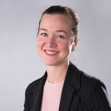 Portrait of a smiling woman with a backdrop that's neutral colored, she's wearing a black blazer over a light pink top.