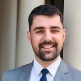 A smiling man in a gray suit and blue tie standing in front of a beige wall.