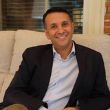 A smiling man in a business suit sitting on a sofa in a room with brick details.