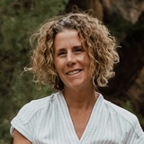 A smiling woman with curly hair, standing outdoors, wearing a light blue v-neck dress.