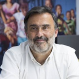 A portrait of a middle-aged man with a beard, wearing a white shirt, smiling gently in an office environment with colorful paintings in the background.