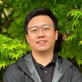 A smiling East Asian man wearing glasses stands in front of lush green foliage.