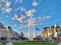 A tall white obelisk monument stands prominently in the center of a bustling city square under a blue sky with scattered clouds.