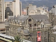 View of a historical building with unique architecture in an urban setting, featuring a modern train passing by and surrounded by taller modern buildings and mountains in the background.