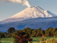 A snow-capped mountain with a plume of smoke rising from its peak, set against a clear sky and foregrounded by lush greenery.