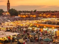 A bustling market square at dusk, with vibrant lights, crowds, and a tall tower in the background under a sunset sky.