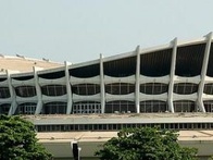 A modern stadium with a distinctive white, curved architecture, surrounded by greenery and a yellow taxi passing by in the foreground.