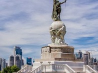 A statue of a warrior on horseback, positioned on a tall pedestal, with a city skyline in the background.