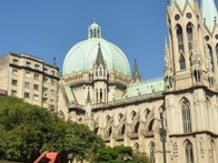 The image shows a large, ornate cathedral with pointed arches and a dome, situated behind a cascading water fountain under a clear blue sky.