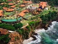 Aerial view of a lush coastal resort with red-roofed buildings next to a rocky cliff and turquoise sea.