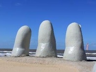 A sculpture of giant fingers buried in sand on a beach with blue sky.