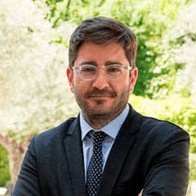 A man in a suit and tie smiling at the camera with trees in the background.