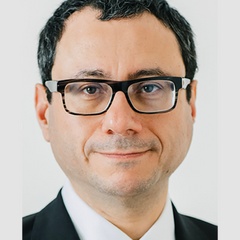 Close-up portrait of a middle-aged man wearing glasses and a black suit, looking directly at the camera.