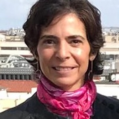 Portrait of a smiling woman with short hair, wearing a black jacket and a pink scarf, against a blurred cityscape background.