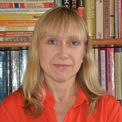 A woman in a red shirt smiling slightly, standing in front of a bookshelf filled with various books.