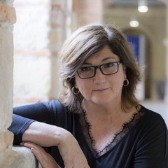 A middle-aged woman with glasses, wearing a black blouse, leans on a stone railing indoors.