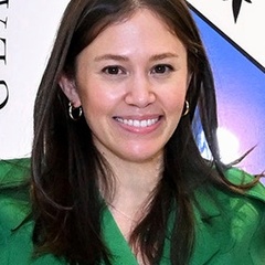 A woman with brown hair wearing a green blouse smiles in front of a promotional banner.