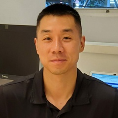 A portrait of a smiling man with short hair, wearing a black polo shirt, standing indoors.
