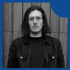 A portrait of a man with long hair and glasses, standing against a wooden background.