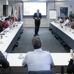 A business meeting with participants seated around a long table listening to a speaker.