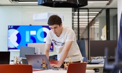 A young man is focused on working at a laptop in a modern workspace surrounded by small robotic models.