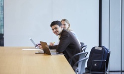 Two young professionals are sitting together at a conference table, working on laptops and looking at the camera.