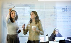 Two women are engaged in a discussion while writing on a glass board, with a third person visible in the background working on a laptop.