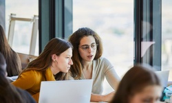 Two women collaborate while working on laptops in a bright, modern workspace.