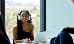 A smiling woman wearing headphones is working on a laptop at a bright and modern office.