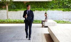 A businessman walks confidently in a modern outdoor space while a woman sits in the background.