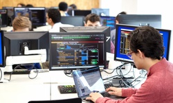 A focused student working on a laptop in a busy computer lab with multiple monitors displaying data.