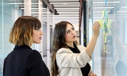 Two women are engaged in a collaborative brainstorming session, reviewing colorful sticky notes on a glass wall in a modern office setting.
